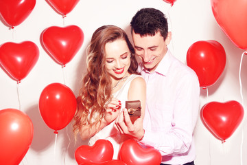 Fototapeta na wymiar Man makes present to his lovely sweetheart girl. Lover's valentine day. Valentine Couple. Boy gives to his girlfriend jewelry. Wedding ring. Propose background red balloons hearts. Happy smile girl.