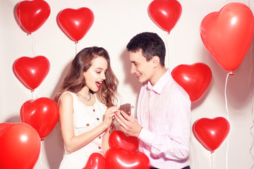 Fototapeta na wymiar Man makes present to his lovely sweetheart girl. Lover's valentine day. Valentine Couple. Boy gives to his girlfriend jewelry. Wedding ring. Propose background red balloons hearts. Happy smile girl.