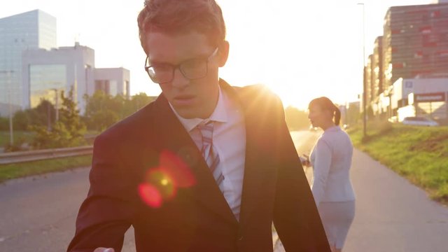 CLOSE UP, LENS FLARE: Businessman and woman fixated on their cell phones bump into each other while walking through downtown Ljubljana at sunset. People concentrating on smart phones on sunny evening.