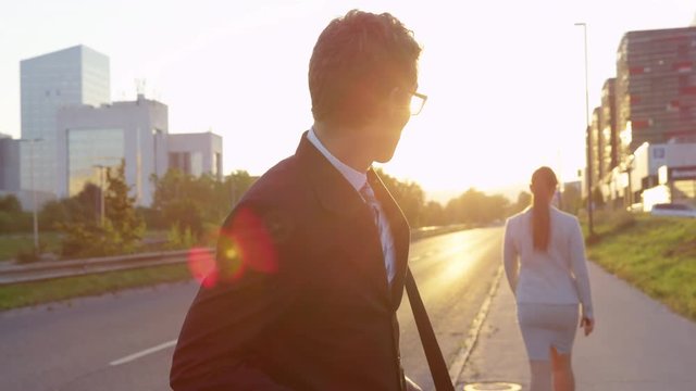 CLOSE UP LENS FLARE: Handsome guy looking at his smartphone sees a gorgeous businesswoman walking to work in the city on a beautiful sunny morning. Businessman stops and stares at gorgeous young woman