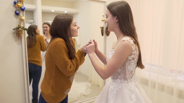 Two brunette girls are in wedding salon.  Girl in dress is always looking in the mirror.They both are very happy and hug because they have chosen the best dress for wedding.