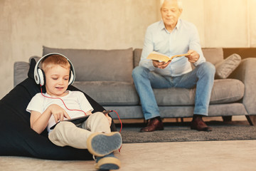 Little boy watching playing on tablet while grandfather reading book