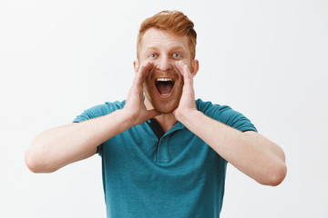 Carefree friendly redhead caucasian man in green polo shirt holding palms near opened mouth while yelling on distance, trying call friend who walking far away, shouting out loud over gray background