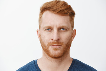 Close-up shot of serious-looking handsome adult european man with red hair and beard staring at camera with focused and determined expression, standing in strict pose over gray background