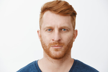 Close-up shot of serious-looking handsome adult european man with red hair and beard staring at camera with focused and determined expression, standing in strict pose over gray background - Powered by Adobe