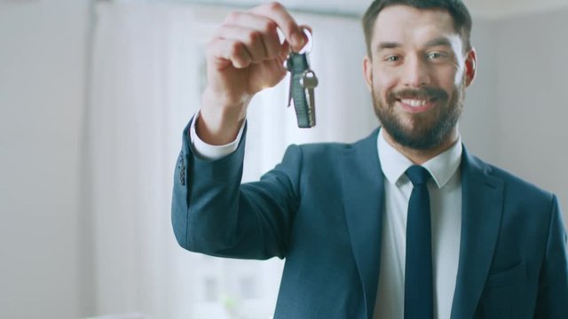 Successful Real Estate Agent in a Suit Smiles and Offers Keys From a New Apartment. Standing in the Middle of Room that Being Renovated. Spacious New House for Sale by Professional Real Estate Broker