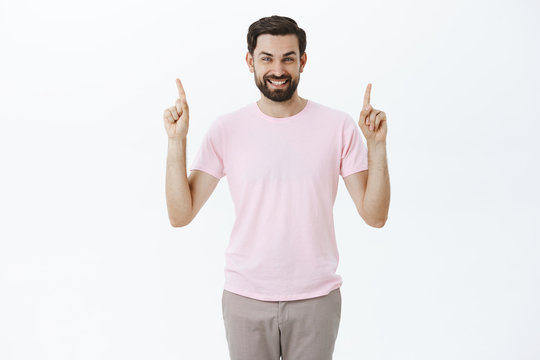 Delighted charismatic happy man with beard and dark hair smiling with broad white grin at camera as showing copy space, pointing up at awesome offer or product, being satified giving recommendation