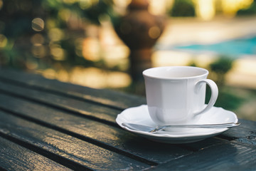 white coffee cup with spoon on wooden table near swimming pool with morning light, in soft focus