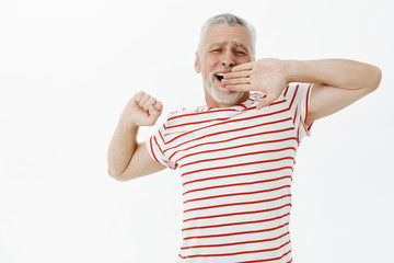Old male pensioner feeling lazy today. Carefree relaxed handsome senior man with beard and grey hair in striped t-shirt stretching and yawning covering opened mouth with palm feeling sleepy and tired