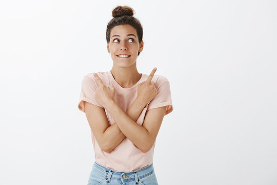 Excited and happy attractive woman with hairbun in t-shirt biting lower lip in anticipation and interest making choice as pointing sideways with crossed arms looking with smile at upper right corner