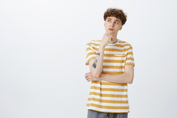 Portrait of thoughtful and concerned young guy thinking up next step holding hand with tattoos on chin looking left focused, making choice or decision posing in striped t-shirt over gray wall