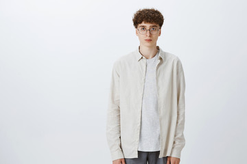Fototapeta premium Studio shot of indifferent unemotional handsome and stylish european hipster boy in glasses with curly haircut standing upright looking at camera with no emotions and feelings, being calm