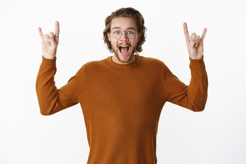 Fototapeta premium Waist-up shot of excited and thrilled overwhelmed attactive bearded man with wavy hair in sweater yelling from amazement and joy showing rock-n-roll gesture buying tickets for favorite band