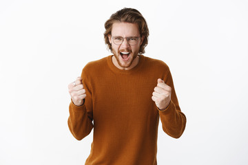 Studio shot of expressive good-looking european bearded guy in sweater and glasses yelling with fure and rage clenching raised fists as cheering and being supportive for favorite team