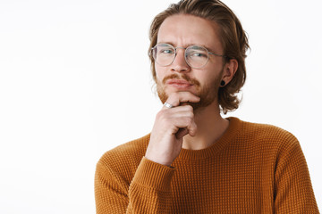 Studio shot of hesitant and unsure good-looking young redhead guy with pierced nose and earrings smirking doubtful holding hand on beard making assumption, thinking doubtful over gray wall