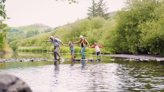 Slow Motion Shot Of Grandparents Helping Grandchildren To Cross River Whilst Hiking In UK Lake District