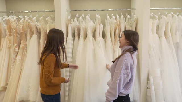 Two brunette girls are in wedding salon. One of them is future bride,another is her best friend. They are choosing dress for fitting.