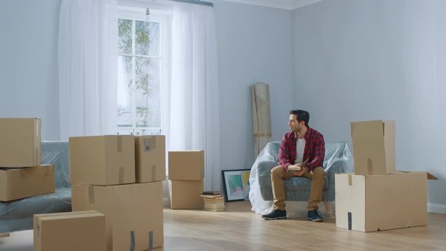 Contemplative Smart Man Reads His Favourite Books While in the Process of Moving Into His New Rented / Purchased Apartment. Unpacked Cardboard Boxes and Covered up Furniture in the Modern Bright Home.