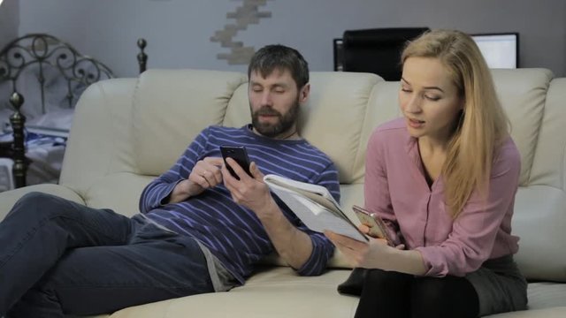 blonde wife on a sofa reads her notes in a notebook and her husband sitting nearby and chatting at the phone
