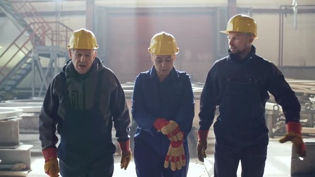 Dolly shot of female engineer in overalls, hard hat and gloves walking through metal fabrication facility and discussing work with male colleagues