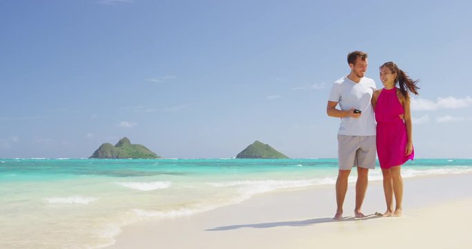 Beach couple using phone app smiling and laughing walking on beach having fun outside under the blue sky . Young Asian woman, Caucasian man. RED EPIC SLOW MOTION