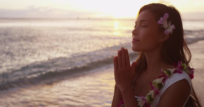 Spiritual - woman giving namaste gesture in yoga meditation on Hawaii wearing traditional Hawaiian flower lei. Meditating serene multicultural young Asian Caucasian woman at ocean beach sunset.