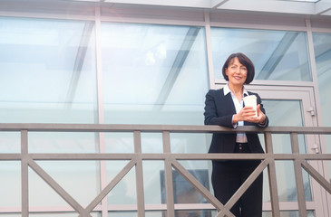 Mature businesswoman having a break in front of an office building, talking on the phone and drinking coffee