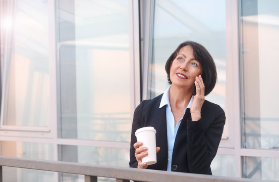 Mature businesswoman having a break in front of an office building, talking on the phone and drinking coffee