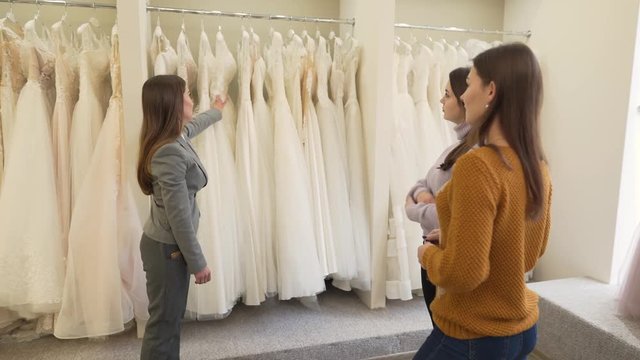 Future bride and her friend are in wedding salon. They want to but wedding dress.They are looking at dresses and listening to shop consultant.