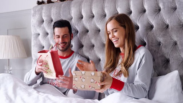 Woman Opening Gift Of Necklace In Bed At Home As Couple Exchange Presents On Christmas Day