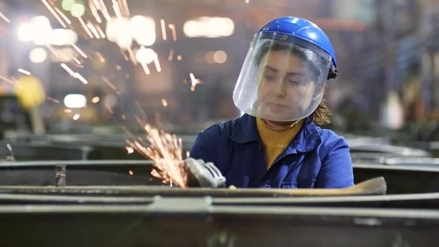 Tracking shot of female worker in safety helmet with face shield using angle grinder tool to polish steel part at metal fabrication facility
