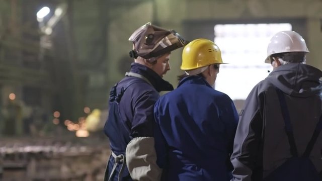 Rear view of female engineer in hard hat walking through metal fabrication facility and discussing work with experienced male worker and young welder in safety uniform