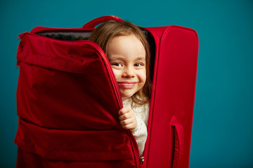 Little girl looks out of red suitcase, portrait of cheerful child on blue isolated background.