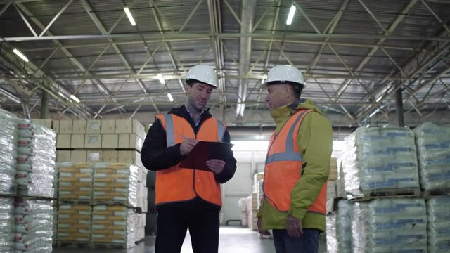 Two employees in workwear and white helmets discussing work in warehouse.