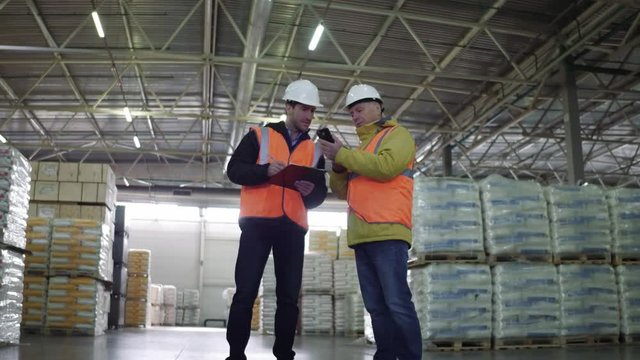 Two employees in workwear and white helmets discussing work in warehouse. Shows the on cellphone.