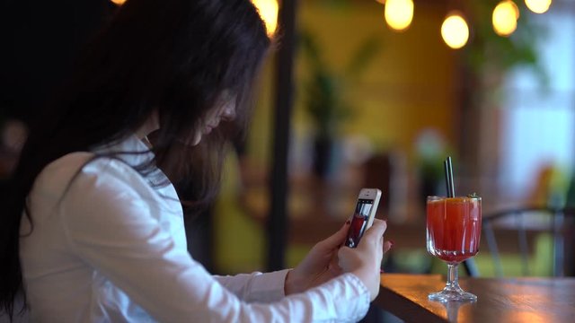 Close-up of woman with phone, taking photos  of glass of orange cocktail with black straws decorated with orange. Back view of lights.