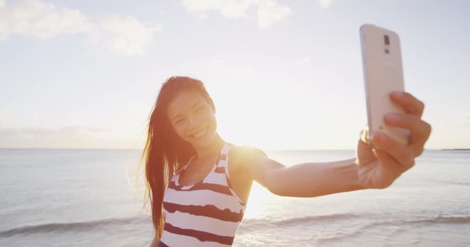 Selfie woman taking photos on phone using smartphone smiling happy on beach. Woman on mobile phone takes self portrait photo pictures outdoors at beach sunset. RED EPIC SLOW MOTION.