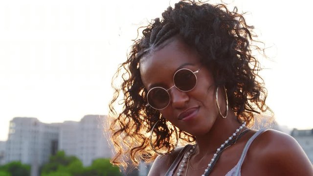 Young black woman wearing sunglasses looking to camera and smiling on a London street at sunset, close up