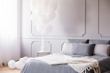 Bunch of white balloons in stylish grey bedroom with king size bed with grey bedding and warm...