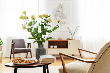 Armchair at table with flowers, books and food in bright apartment interior with windows. Real photo