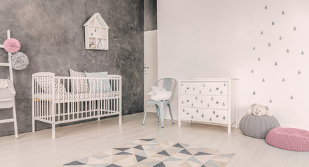 White cradle next to chair and cabinet in grey baby's bedroom interior with pink pouf. Real photo