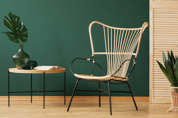 Wicker chair next to wooden table with black glass vase and monstera leaf, copy space on the empty green wal