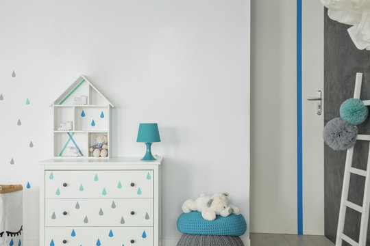Blue lamp on white cabinet next to teddy bear on pouf in child's room interior. Real photo