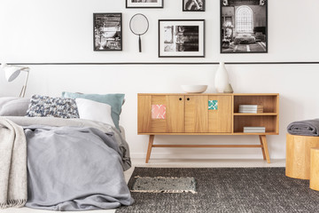 Wooden cupboard next to bed with sheets in grey bedroom interior with carpet and gallery. Real photo