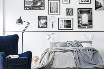 Lamp next to armchair in white bedroom interior with gallery above bed with sheets. Real photo