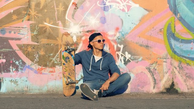 Male teenager is sitting near a graffiti wall with his skateboard