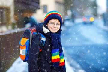 Little school kid boy of elementary class walking to school during snowfall. Happy healthy child with glasses having fun and playing with snow. With backpack or satchel in colorful winter clothes.