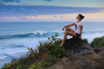 Girl posing sitting sideways on a stone cliff against the background of the blue sea and sky