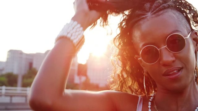 Close up of young black woman wearing sunglasses plying with her hair, looking to camera on a London street at the golden hour