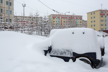 Cars covered with snow after a snowfall. Around the snowdrifts and a lot of snow on the machines. In the background multicolored panel buildings. Cold winter weather. Magadan, Siberia, Far East Russia